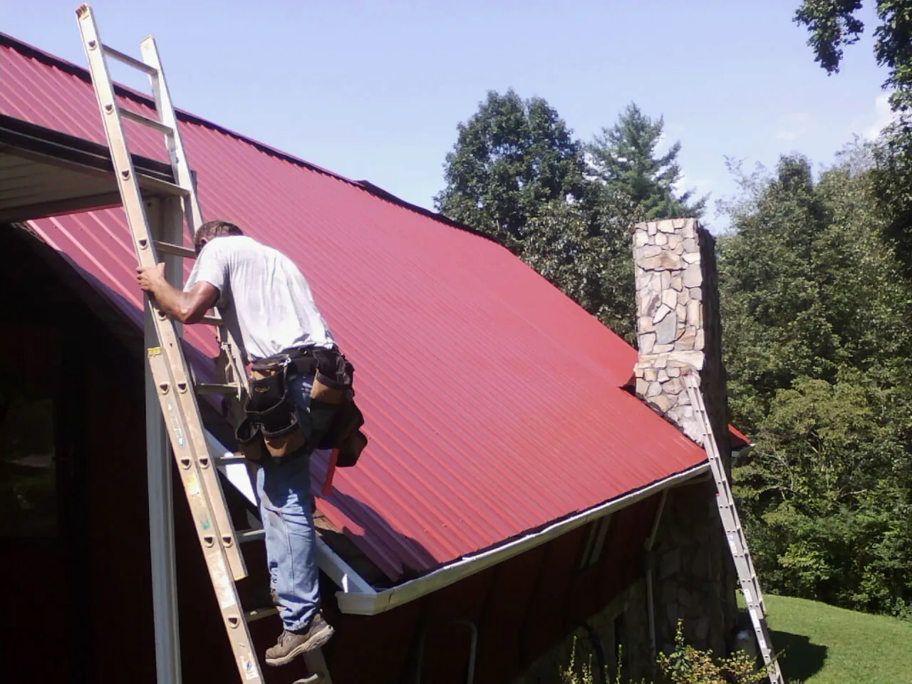 Roofer on a ladder beside a red metal roof and stone chimney — Soffit & Fascia Repair by Roofing Brothers in Netawaka